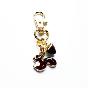 Peut inclure: Un porte-clés en métal doré avec un fermoir mousqueton et deux breloques. Une breloque représente un écureuil en émail marron foncé et blanc, l'autre est un gland marron foncé avec un capuchon doré texturé. Les breloques sont fixées par des anneaux.