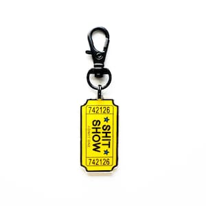 May include: A yellow rectangular keyring with the text "SHIT SHOW" and the number "742126" printed on it. The keyring has a black metal clasp and ring.