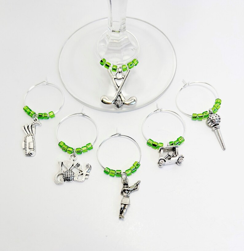 May include: Set of six green and silver golf-themed wine glass charms. Each charm features a different golf-related design, including golf clubs, a golf ball, a golf cart, a golfer, and a golf bag.