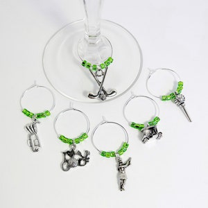 May include: Set of six green and silver wine glass charms featuring golf-themed charms, including a golf cart, golf clubs, a golfer, and a golf ball.
