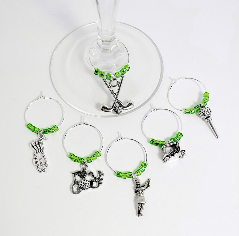 May include: Set of six green and silver wine glass charms featuring golf-themed charms, including a golf cart, golf clubs, a golfer, and a golf ball.