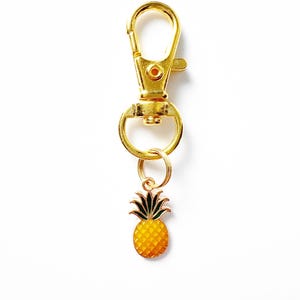 May include: Gold-colored keychain with a lobster clasp and a small pineapple charm. The pineapple charm is yellow with green leaves and a textured surface. The keychain is on a white background.