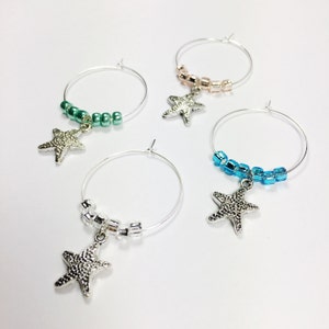 May include: Four silver wine glass charms with star-shaped pendants and colorful beaded strands. The charms are in green, blue, pink, and silver.