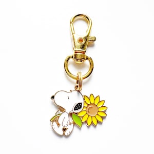 May include: A gold-colored metal keychain featuring a white cartoon character with black ears and a black spot, holding a sunflower. The sunflower has yellow petals and a brown center. The keychain has a lobster clasp.