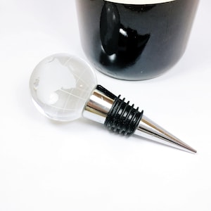May include: A silver and black wine bottle stopper with a clear glass globe top featuring a raised world map design.