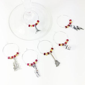 May include: A set of six silver-toned metal wine glass charms, each featuring a circular hoop decorated with small red and gold beads. The charms include charms shaped like a castle, a lightning bolt, a cauldron, a broom, and a deer.
