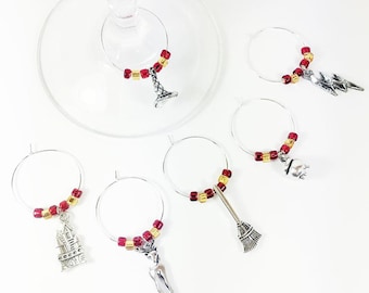 May include: A set of six silver-toned metal wine glass charms, each featuring a circular hoop decorated with small red and gold beads. The charms include charms shaped like a castle, a lightning bolt, a cauldron, a broom, and a deer.