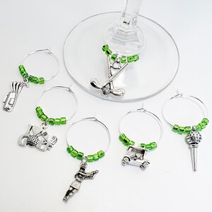 May include: Set of six green and silver golf-themed wine glass charms. Each charm features a different golf-related design, including a golf bag, golf ball, golf club, golf cart, and a golfer.