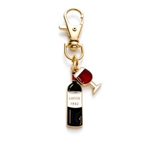 May include: A gold-toned metal keychain featuring two enamel charms: a wine bottle with a black and white label reading LAFITE 1982 and a red wine glass. The charms are attached to a gold-toned lobster clasp by a small circular jump ring.