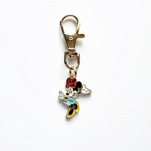 May include: A gold-colored metal keychain with a clasp and a charm featuring Minnie Mouse. The Minnie Mouse charm is in color, with a red bow, black ears, a blue top, yellow shoes, and white gloves.