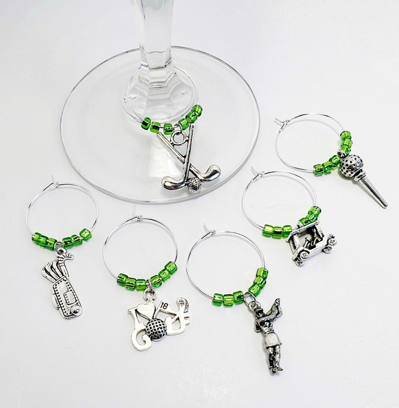 May include: Six green and silver wine glass charms with golf themed charms. The charms include a golf club, a golf cart, a golfer, a golf bag, a golf ball, and a golf tee.