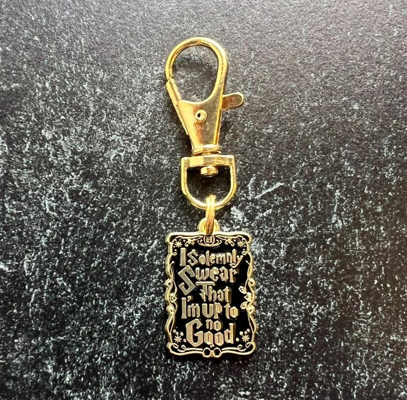 May include: Gold-tone keychain with a black rectangular charm. The charm displays the phrase "I Solemnly Swear That I'm Up to No Good" in white, with floral embellishments. The keychain has a lobster clasp for attachment.