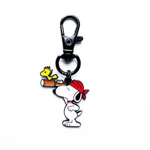 May include: A metal keychain featuring a white dog character wearing a red bandana and looking through a brown telescope, with a small yellow bird perched on the telescope. The charm is attached to a black metal lobster claw clasp.