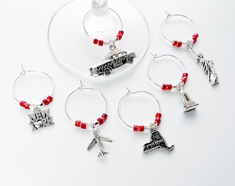 May include: Set of six red and silver wine glass charms featuring New York City themed charms, including a taxi, the Statue of Liberty, the Empire State Building, a plane, the state of New York, and a charm that says "New York" with a heart.