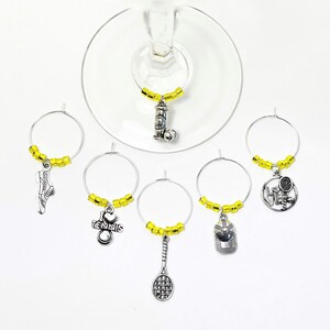 May include: A set of six silver-toned metal wine glass charms featuring tennis-themed pendants including a racket, a shoe, a cap, a ball canister, and a heart with the text I LOVE TENNIS, each accented with small yellow beads on a circular wire hoop.