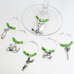 May include: Set of six green and silver golf-themed wine glass charms. Each charm features a different golf-related design, including a golf bag, golf ball, golf club, golf cart, and a golfer.