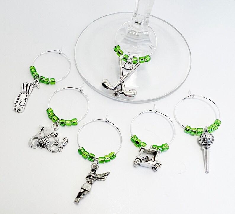 May include: Set of six green and silver golf-themed wine glass charms. Each charm features a different golf-related design, including a golf bag, golf ball, golf club, golf cart, and a golfer.