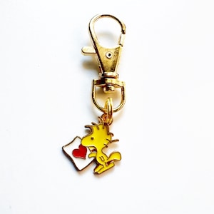 May include: A gold-colored metal keychain with a clasp. The charm is a yellow cartoon bird holding a white envelope with a red heart. The keychain is on a white background.