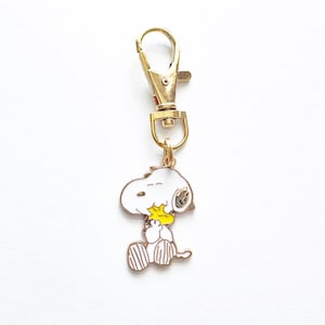 May include: Gold-tone keychain featuring a white cartoon dog holding a yellow bird. The keychain has a gold-tone lobster clasp.