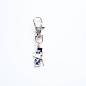 May include: A Snoopy keychain charm in a formal outfit, including a top hat and cane. The charm is attached to a silver-colored metal clasp. The Snoopy figure is white with black accents and a blue suit.