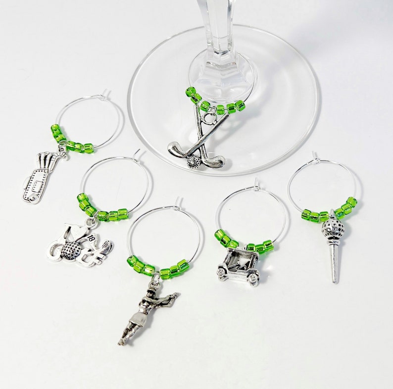 May include: Set of six green and silver golf-themed wine glass charms. Each charm features a different golf-related design, including a golf club, golf ball, golf cart, and a golfer.
