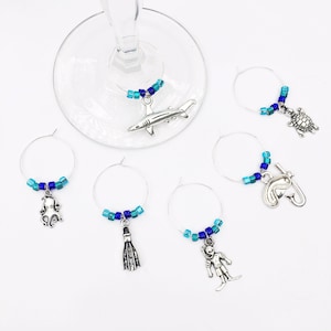 May include: Set of six silver wine glass charms with blue and turquoise beads. Each charm features a different sea creature: shark, turtle, octopus, scuba diver, flipper, and a fish with a snorkel.