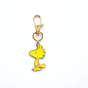May include: A gold-toned metal keyring featuring a yellow enamel charm of the character Woodstock. The charm is attached to a gold-coloured lobster claw clasp by a small circular jump ring. The character is depicted in profile facing left.