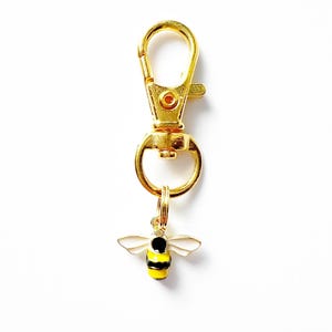 May include: A gold-colored keychain with a lobster clasp and a small bee charm. The bee charm has a black head, yellow and black striped body, and white wings. The keychain is on a white background.