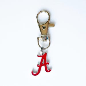 May include: A silver-colored metal keychain clip with a red letter "A" charm. The charm is a stylized script letter with a glossy finish. The keychain clip is attached to a small metal ring.