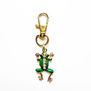 May include: A gold-colored metal keychain with a green enamel frog charm. The frog has rhinestone accents and gold-colored legs and arms. The keychain clasp is also gold-colored.