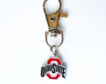 May include: A silver-coloured keychain clip with a swivel base and a charm. The charm features the Ohio State logo in red, black, and silver, with the words "OHIO STATE" in a bold font.