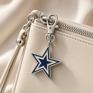 May include: A cream-colored pouch featuring a silver-toned metal zipper and a detachable star-shaped charm. The charm is navy blue with a white border and a silver-toned metal outline, attached to the zipper pull with a silver lobster clasp.