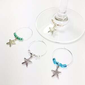 May include: Four silver starfish wine glass charms with different colored beads. The charms are hanging from a silver wire ring. The beads are green, white, blue, and pink.