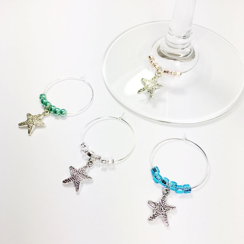 May include: Four silver starfish wine glass charms with different colored beads. The charms are hanging from a silver wire ring. The beads are green, white, blue, and pink.