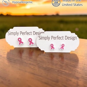 May include: Two pairs of pink ribbon stud earrings displayed on white Simply Perfect Design cards. The earrings feature a pink awareness ribbon shape with a white border. Icons indicate the items are handcrafted, hypoallergenic, and made in the USA.