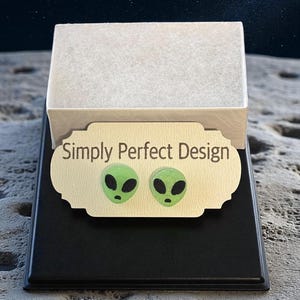 Op de afbeelding: Een zwarte displaystandaard met een goudkleurige plaat met de tekst "Simply Perfect Design". Twee groene, buitenaardse oorbellen worden op de plaat getoond. De achtergrond is een gestructureerd oppervlak met een sterrenhemel.