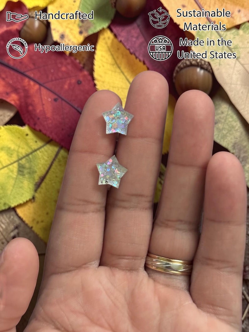 Op de afbeelding: Twee stervormige oorbellen met iriserende glitter worden in een hand gehouden. De oorbellen zijn handgemaakt, hypoallergeen en gemaakt in de VS met duurzame materialen. De achtergrond bevat herfstbladeren en eikels.