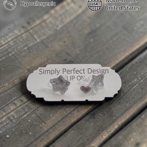 Op de afbeelding: Een displaykaart met de tekst "Simply Perfect Design" toont twee stervormige oorbellen en &eacute;&eacute;n hartvormige oorbel. De oorbellen zijn transparant met glitter. De kaart heeft ook tekst met "Handcrafted", "Hypoallergenic", "Sustainable Materials" en "Made in the United States".