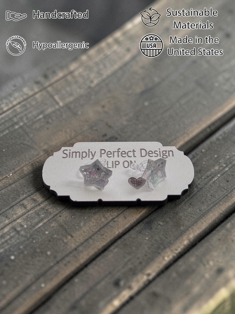 Op de afbeelding: Een displaykaart met de tekst "Simply Perfect Design" toont twee stervormige oorbellen en &eacute;&eacute;n hartvormige oorbel. De oorbellen zijn transparant met glitter. De kaart heeft ook tekst met "Handcrafted", "Hypoallergenic", "Sustainable Materials" en "Made in the United States".
