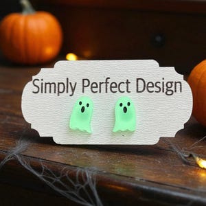 Może przedstawiać: Para jasnozielonych kolczyków w kształcie ducha na białej karcie z napisem "Simply Perfect Design". Kolczyki mają czarne oczy i uśmiechnięte usta. Tło obejmuje dynie i drewnianą powierzchnię.