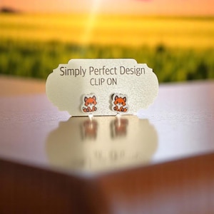 Könnte beinhalten: Clip-Ohrringe mit einem Fuchsmotiv. Die Ohrringe sind orange und weiß, präsentiert auf einer weißen Karte mit dem Text "Simply Perfect Design CLIP ON". Der Hintergrund ist ein verschwommener Sonnenuntergang.