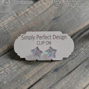 Op de afbeelding: Stervormige clip-on oorbellen met iriserende glitter, gepresenteerd op een witte kaart. De kaart leest "Simply Perfect Design" en "CLIP ON". De afbeelding bevat ook tekst die aangeeft dat de oorbellen handgemaakt, hypoallergeen en gemaakt in de VS met duurzame materialen zijn.
