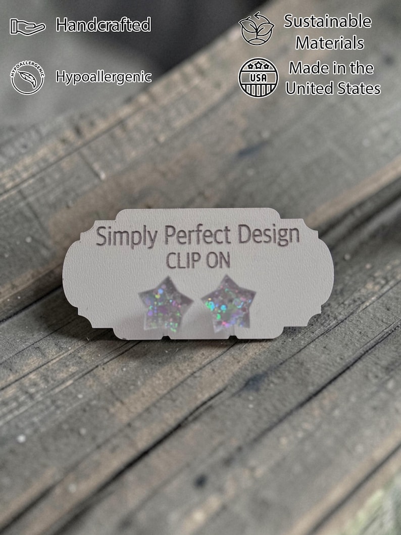 Op de afbeelding: Stervormige clip-on oorbellen met iriserende glitter, gepresenteerd op een witte kaart. De kaart leest "Simply Perfect Design" en "CLIP ON". De afbeelding bevat ook tekst die aangeeft dat de oorbellen handgemaakt, hypoallergeen en gemaakt in de VS met duurzame materialen zijn.