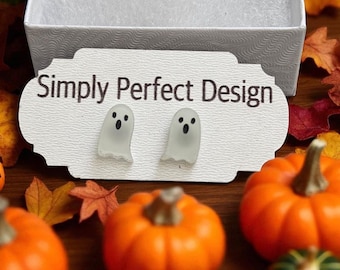 May include: A pair of ghost-shaped earrings with black eyes and mouths, displayed on a white card with the text "Simply Perfect Design." The earrings are set against a backdrop of orange pumpkins and autumn leaves, suggesting a Halloween theme.