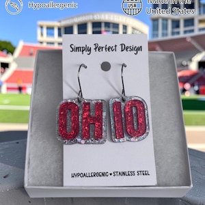 May include: A pair of dangle earrings on a white display card labeled Simply Perfect Design. The earrings feature the word OHIO in red glitter letters set against a clear, silver-flecked background. They hang from dark metal hooks and are marked as hypoallergenic stainless steel.