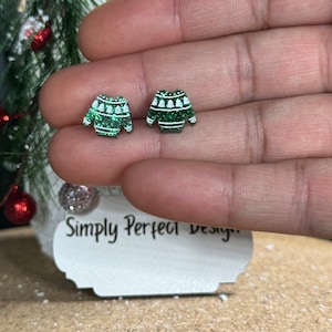 May include: A pair of stud earrings shaped like miniature green jumpers with white glittery accents. The jumpers feature a horizontal striped pattern with small white tree motifs across the chest area. The earrings are displayed on a white card.