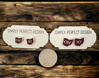 May include: Two pairs of stud earrings shaped like the state of Ohio, featuring a dark red finish with white text reading OH and IO. The earrings are displayed on two cream-colored cards labeled SIMPLY PERFECT DESIGN, positioned above a US quarter.