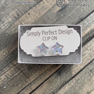 Op de afbeelding: Stervormige clip-on oorbellen met iriserende glitter, gepresenteerd in een witte doos. De doos heeft de tekst "Simply Perfect Design" en "CLIP ON". De afbeelding geeft ook aan dat de oorbellen handgemaakt, hypoallergeen, gemaakt van duurzame materialen en gemaakt in de VS zijn.