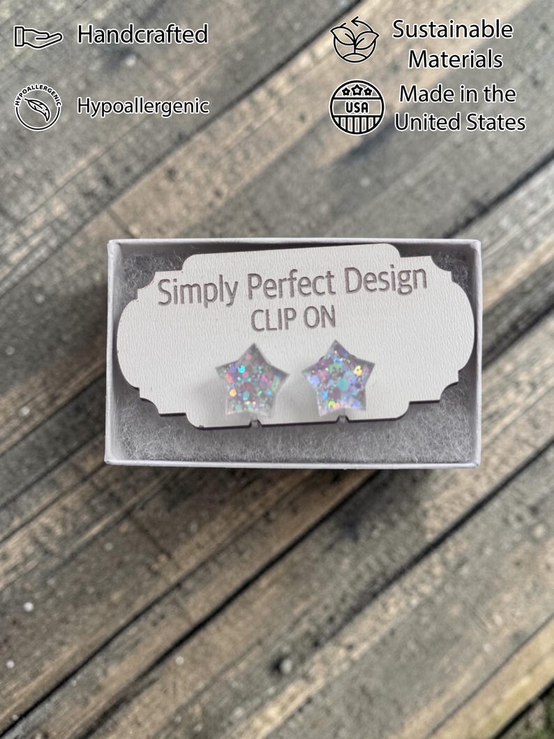 Op de afbeelding: Stervormige clip-on oorbellen met iriserende glitter, gepresenteerd in een witte doos. De doos heeft de tekst "Simply Perfect Design" en "CLIP ON". De afbeelding geeft ook aan dat de oorbellen handgemaakt, hypoallergeen, gemaakt van duurzame materialen en gemaakt in de VS zijn.