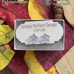 Op de afbeelding: Stervormige clip-on oorbellen met glitter, gepresenteerd in een kleine witte doos. De doos ligt op een bed van kleurrijke herfstbladeren. De tekst "Simply Perfect Design" en "CLIP ON" zijn zichtbaar op de doos.
